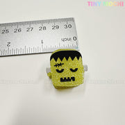 A small glittery yellow Frankenstein's monster from the Kawaii Halloween Hair Claw Clips – Set of 8 Spooky-Cute Designs is shown next to a ruler (about 3 cm tall). Tiny Mochi Gift Shop Houston branding appears in pastel in the corner.