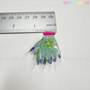 A glittery green monster paw-shaped pin with purple claws and yellow spots, about 4cm long, sits below a ruler for scale. Fun addition to the Kawaii Halloween Hair Claw Clips set by Tiny Mochi Gift Shop Houston: Cute accessories & more!.