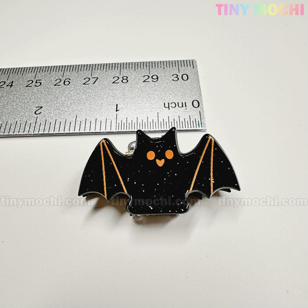 A black bat-shaped enamel pin with orange eyes is pictured near a metal ruler (about 2 inches), on a white background with "Tiny Mochi Gift Shop Houston" at the top, alongside Kawaii Halloween Hair Claw Clips – Set of 8 Spooky-Cute Designs.