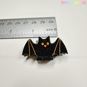 A black bat-shaped enamel pin with orange eyes is pictured near a metal ruler (about 2 inches), on a white background with "Tiny Mochi Gift Shop Houston" at the top, alongside Kawaii Halloween Hair Claw Clips – Set of 8 Spooky-Cute Designs.