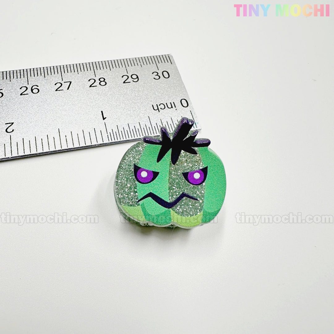 A small glittery green pumpkin charm with a frown and purple eyes sits by a ruler for scale. About 3cm wide, it pairs perfectly with Kawaii Halloween Mini Claw Clips from Tiny Mochi Gift Shop Houston: Cute Hair clips and Accessories.