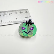 A small glittery green pumpkin charm with a frown and purple eyes sits by a ruler for scale. About 3cm wide, it pairs perfectly with Kawaii Halloween Mini Claw Clips from Tiny Mochi Gift Shop Houston: Cute Hair clips and Accessories.