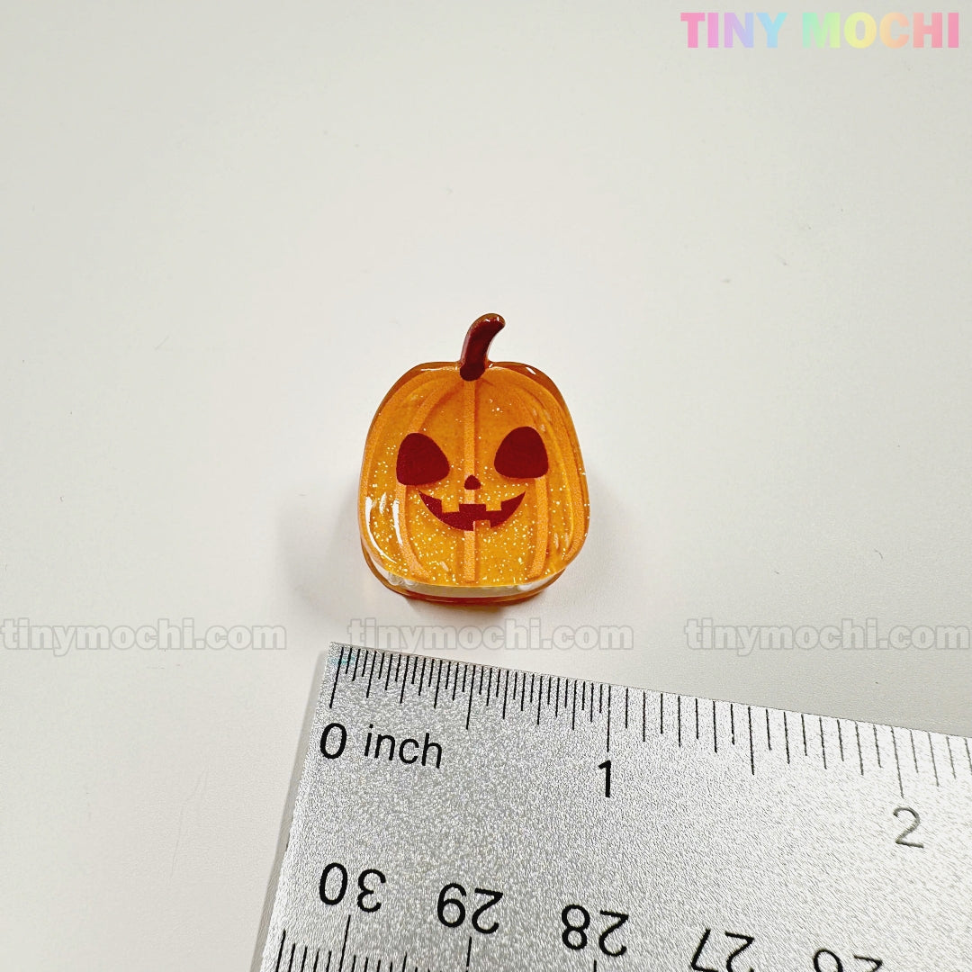 A small, orange pumpkin charm with a jack-o-lantern face sits by a silver ruler on a white surface—perfect for your collection of Kawaii Halloween Mini Claw Clips from Tiny Mochi Gift Shop Houston. Pastel "TINYMochi" appears at the top right.