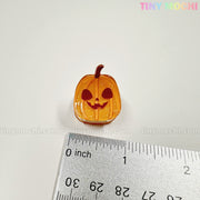 A small, orange pumpkin charm with a jack-o-lantern face sits by a silver ruler on a white surface—perfect for your collection of Kawaii Halloween Mini Claw Clips from Tiny Mochi Gift Shop Houston. Pastel "TINYMochi" appears at the top right.