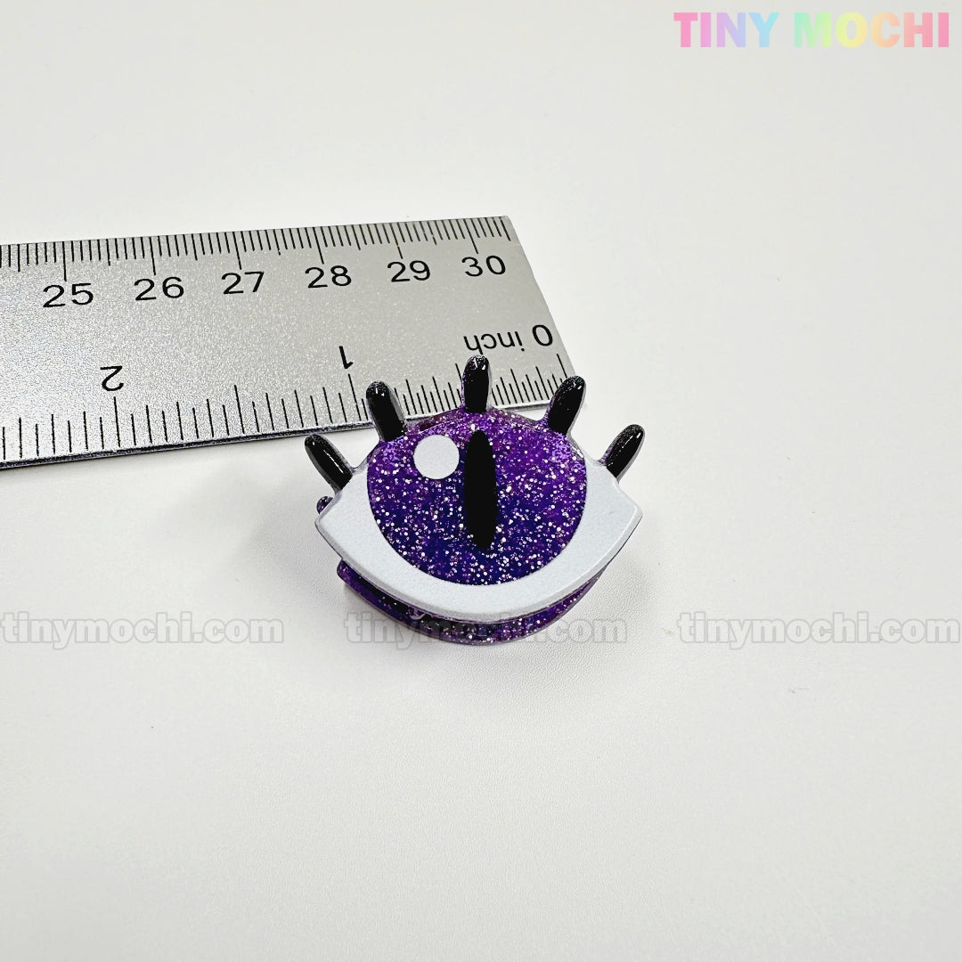 A small, glittery purple eye-shaped pin with black lashes sits by a ruler—just over 1 inch wide. Pairs well with Kawaii Halloween Mini Claw Clips from Tiny Mochi Gift Shop Houston. "TINY MOCHI" is faintly visible on the white background.
