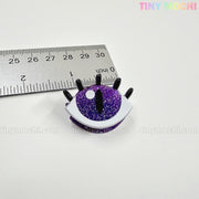 A small, glittery purple eye-shaped pin with black lashes sits by a ruler—just over 1 inch wide. Pairs well with Kawaii Halloween Mini Claw Clips from Tiny Mochi Gift Shop Houston. "TINY MOCHI" is faintly visible on the white background.