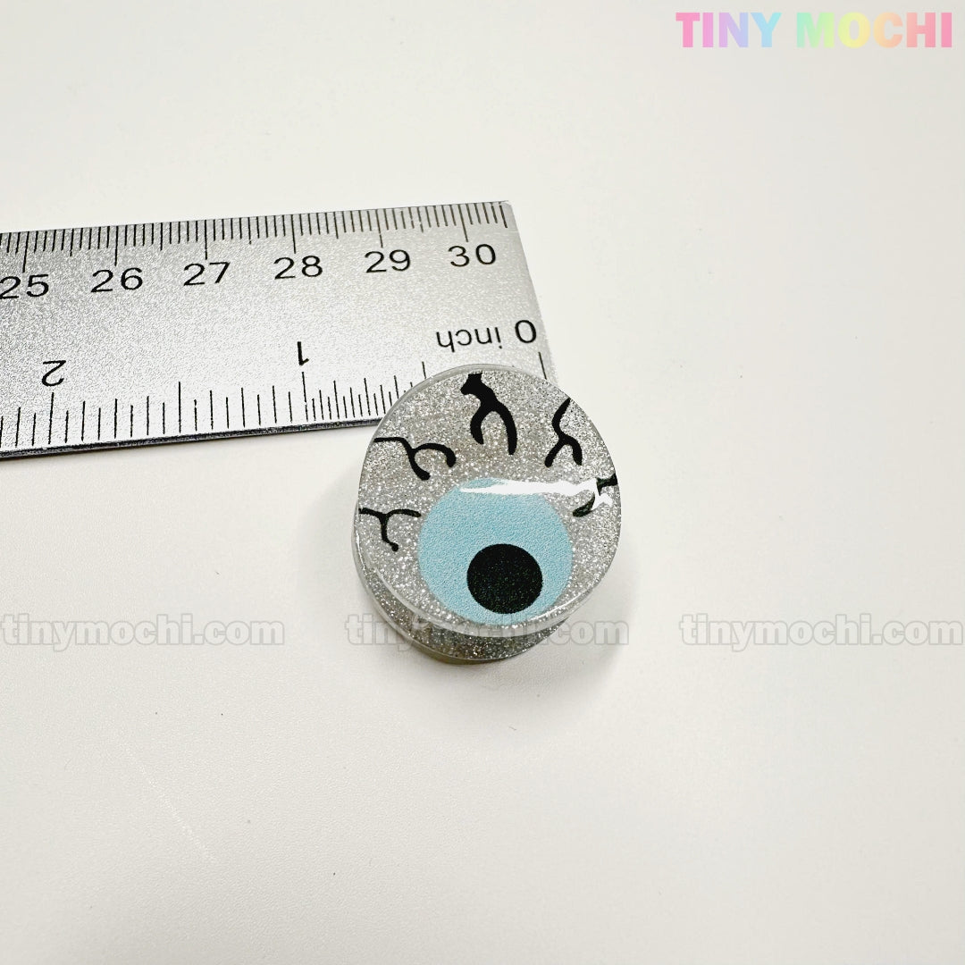 A round, glittery pin shaped like a blue eyeball with black veins is shown next to a ruler (about 1 inch wide). Perfect for pairing with Kawaii Halloween Mini Claw Clips by Tiny Mochi Gift Shop Houston. White background, TINYMochi in corners.