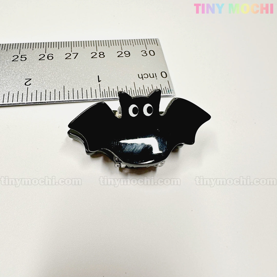 A shiny black bat-shaped Kawaii Halloween Mini Claw Clip by Tiny Mochi Gift Shop Houston sits next to a silver ruler on a white background with "TINYMochi.com" faintly visible. Perfect spooky cute hair accessory for Halloween!.
