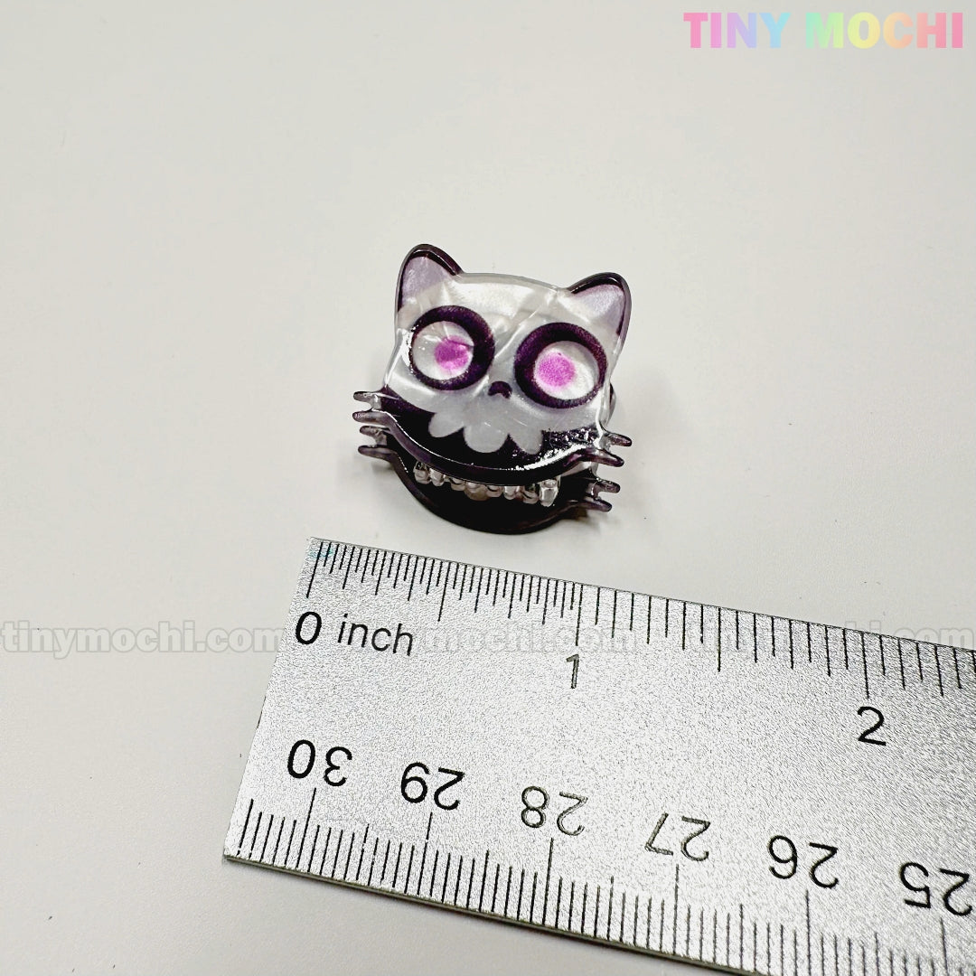 A small, purple cat-shaped keycap with a skull face sits by a metal ruler measuring about 1 inch wide. "TINY MOCHI" appears in pastels—perfect for fans of the Kawaii Halloween Mini Claw Clips from Tiny Mochi Gift Shop Houston.