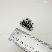 A small, glittery spiderweb pin sits by a metal ruler, ready to style with Kawaii Halloween Mini Claw Clips from Tiny Mochi Gift Shop Houston. "TINY MOCHI" in pastel colors decorates the white background’s top right corner.