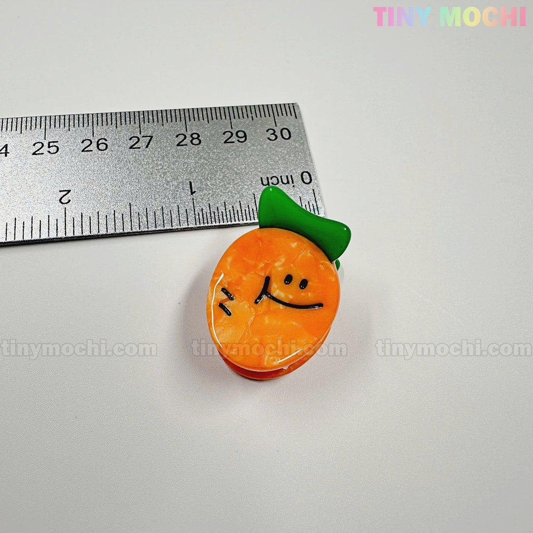 A cute orange veggie charm from the Kawaii Vegetable Hair Clip Set (8 pieces) by Tiny Mochi Gift Shop Houston is shown by a ruler for scale. Ideal for Halloween hair accessories, with tinymochi.com faintly visible in the background.