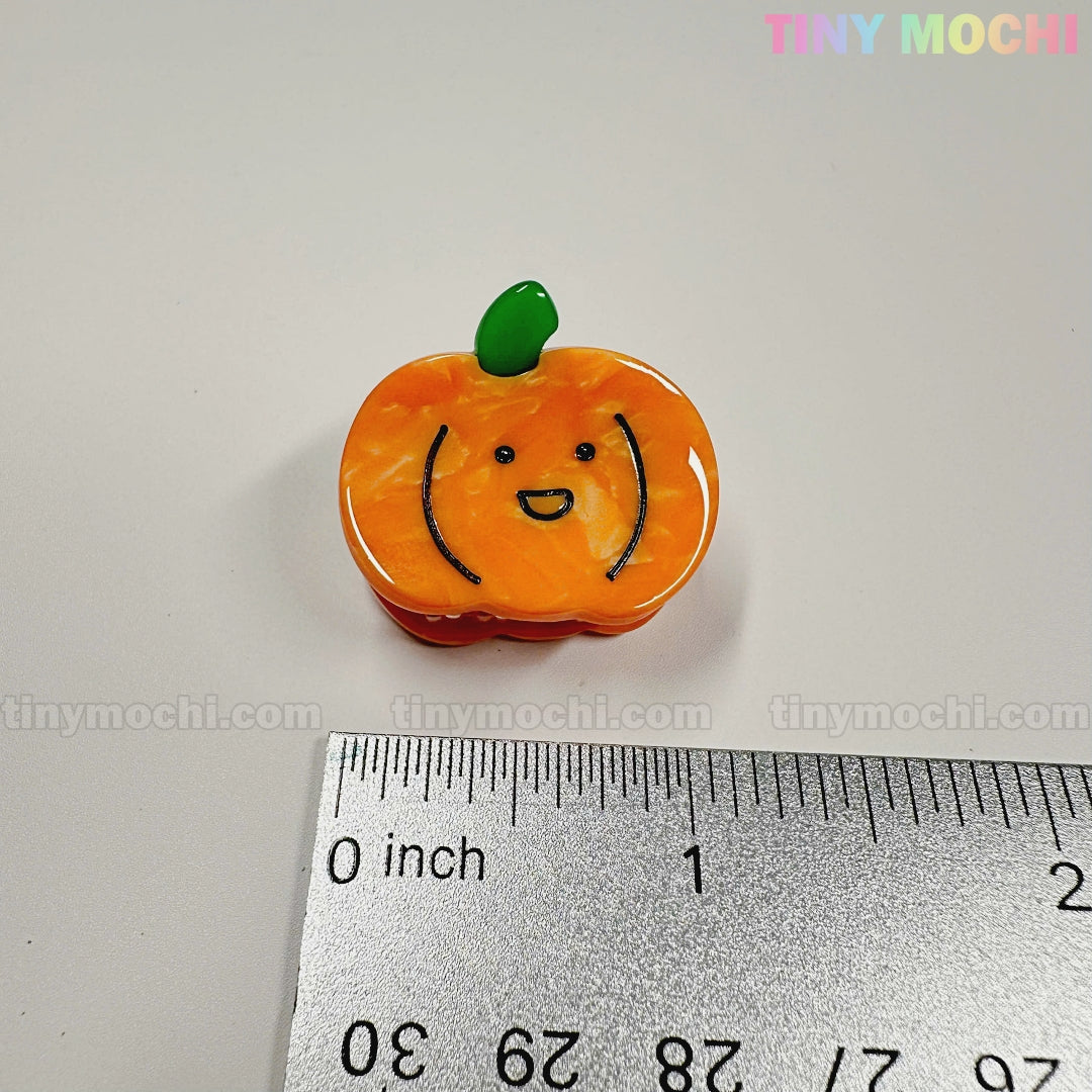 A cute, orange pumpkin-shaped charm with a smiling face from the Kawaii Vegetable Hair Clip Set – 8 Pieces by Tiny Mochi Gift Shop Houston sits on a white surface near a ruler—perfect for kawaii Halloween hair clips.