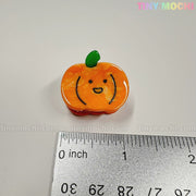 A cute, orange pumpkin-shaped charm with a smiling face from the Kawaii Vegetable Hair Clip Set – 8 Pieces by Tiny Mochi Gift Shop Houston sits on a white surface near a ruler—perfect for kawaii Halloween hair clips.