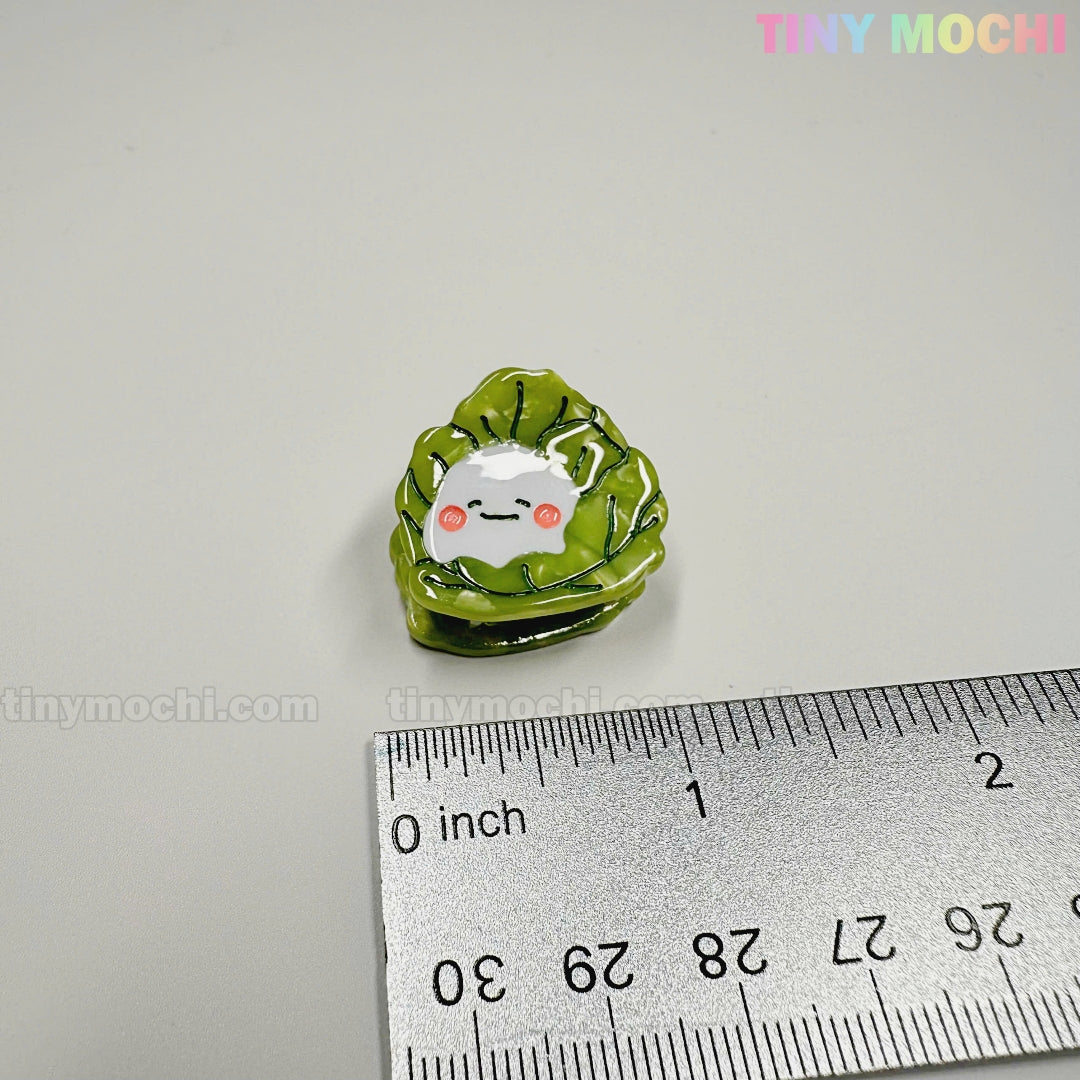 A small enamel pin shaped like a smiling white dumpling wrapped in green cabbage leaves sits by a ruler. TINY MOCHI is printed in colorful letters, making it as charming as the Kawaii Vegetable Hair Clip Set from Tiny Mochi Gift Shop Houston.