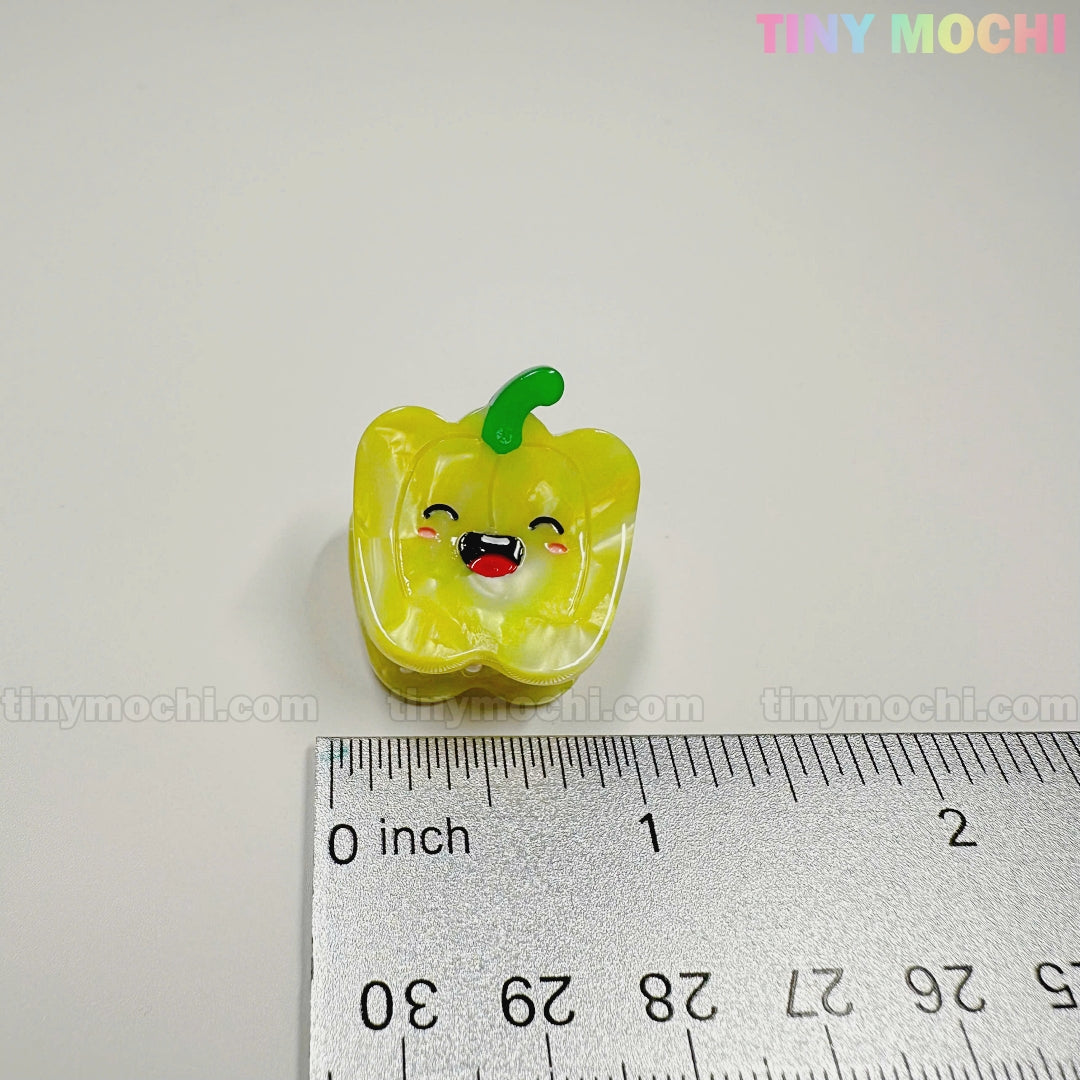 A small, yellow, bell pepper-shaped hair clip from the Kawaii Vegetable Hair Clip Set – 8 Pieces (Tiny Mochi Gift Shop Houston) is shown beside a ruler for scale, with tinymochi.com in the corners for a cute finishing touch.