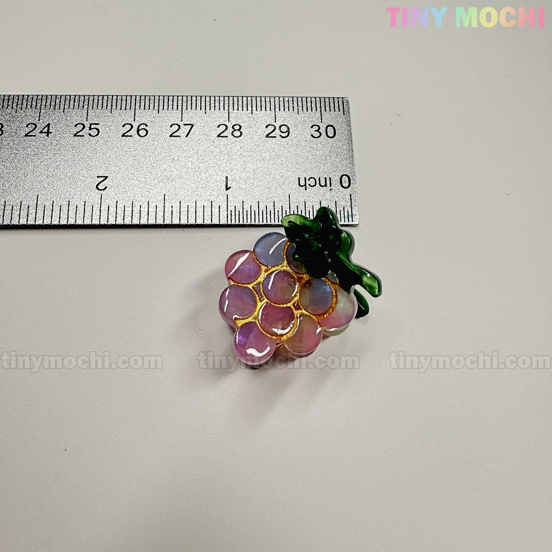 A small, shiny grape-shaped charm with gold outlines and green leaves sits by a ruler showing it's ~3 cm wide. "Fruity Friends Hair Clip Set" by Tiny Mochi Gift Shop Houston—ideal for kawaii hair accessories fans!.