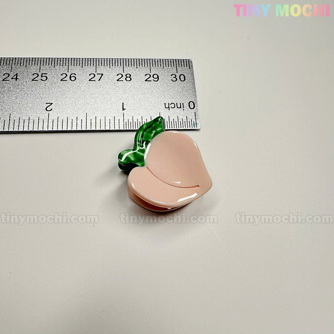 A small, peach-shaped pin from the Fruity Friends Hair Clip Set by Tiny Mochi Gift Shop Houston is shown next to a ruler (about 1 inch wide). The light background has a faint tinymochi.com watermark. Perfect for cute, fruity hair accessories!.