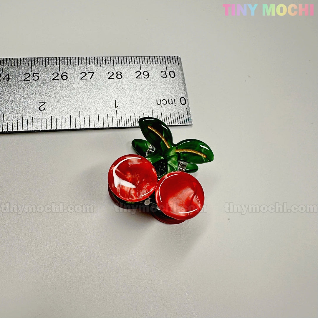 A red cherry-shaped hair clip with green leaves from the Fruity Friends Hair Clip Set by Tiny Mochi Gift Shop Houston sits next to a metal ruler, measuring about 4 cm wide. The Tiny Mochi logo appears in the top right corner.