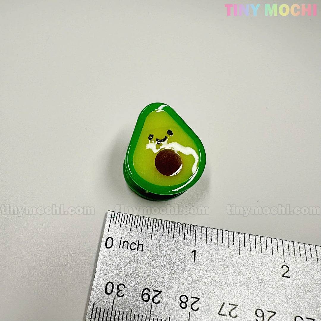 A cute, 1-inch avocado-shaped hair clip from the Fruity Friends Hair Clip Set by Tiny Mochi Gift Shop Houston sits on a white surface next to a ruler. Perfect kawaii accessory for kids' hair or collections! "TINY MOCHI" appears in the corners.