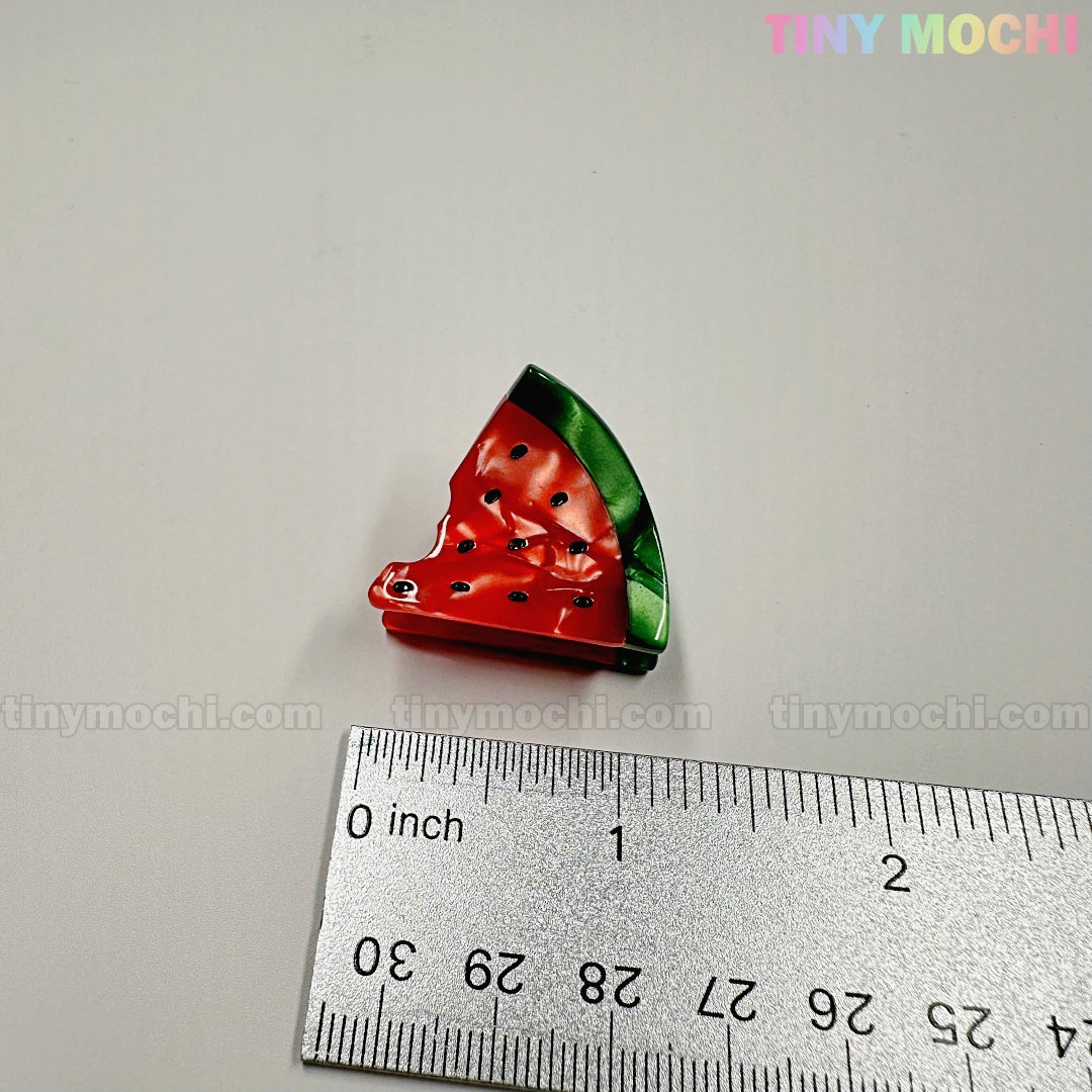 A Fruity Friends Hair Clip from Tiny Mochi Gift Shop Houston, shaped like a bitten watermelon, sits by a ruler on a white surface. TINY MOCHI and tinymochi.com are shown. Ideal for fans of cute hair clips and kawaii accessories!.