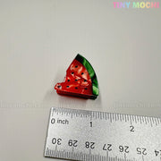 A Fruity Friends Hair Clip from Tiny Mochi Gift Shop Houston, shaped like a bitten watermelon, sits by a ruler on a white surface. TINY MOCHI and tinymochi.com are shown. Ideal for fans of cute hair clips and kawaii accessories!.