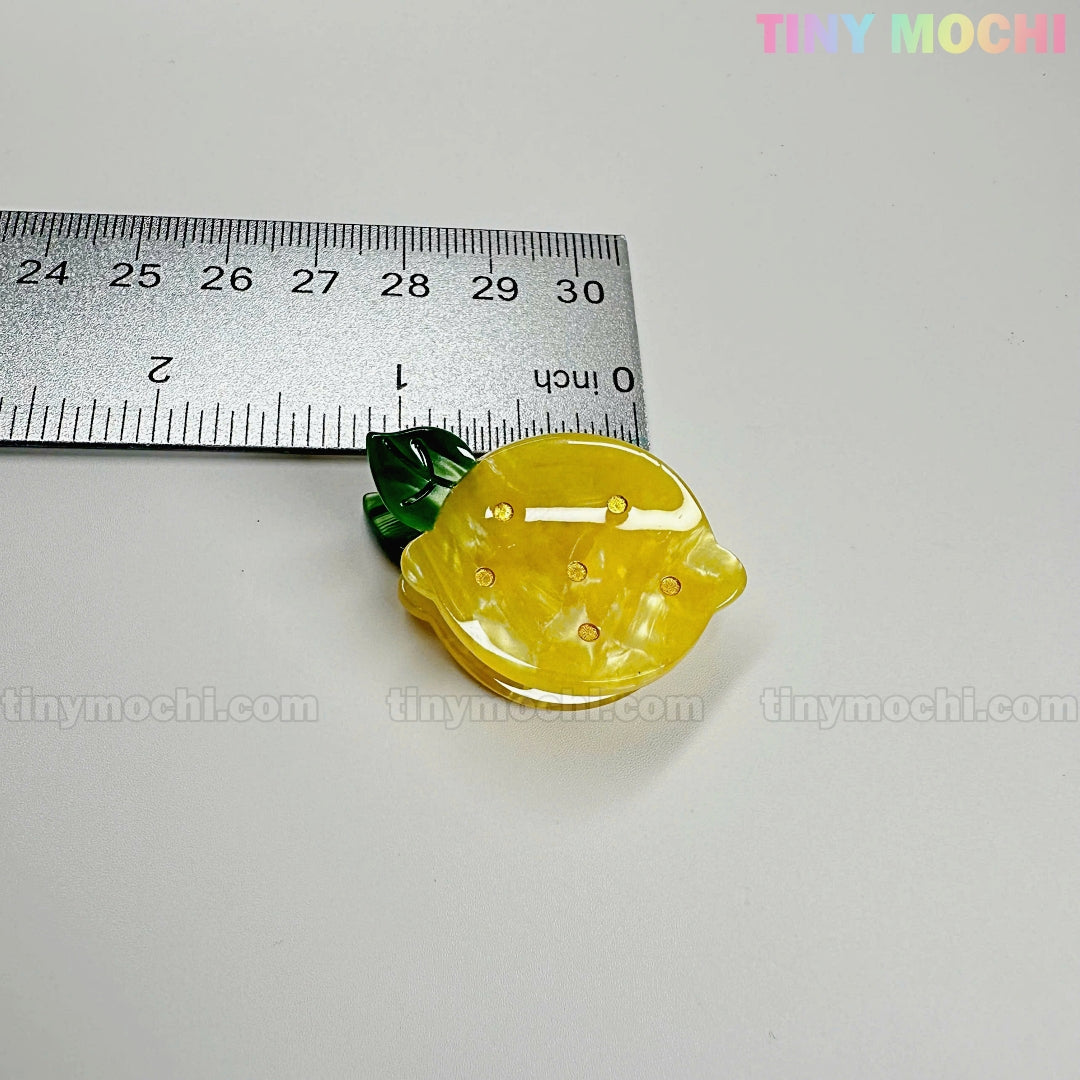 A yellow lemon-shaped acrylic charm with green leaves from the Fruity Friends Hair Clip Set by Tiny Mochi Gift Shop Houston is shown next to a ruler (1.5 in) on a white surface. "tiny mochi" appears faintly in the image.